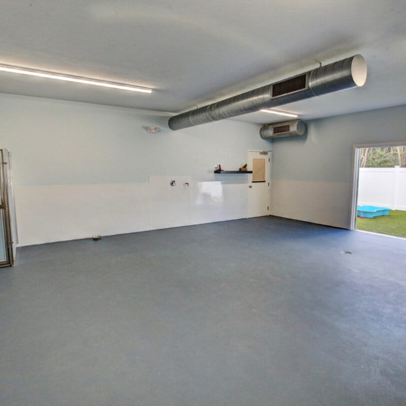 Spacious indoor playroom with clean, non-slip flooring, large ventilation ducts, and a wide door leading to an outdoor grass area with a kiddie pool.