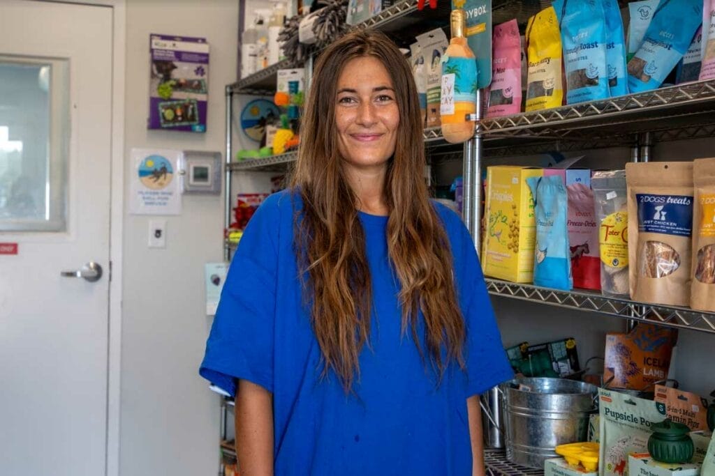 Female team member standing and smiling inside a pet supply room, with shelves behind her stocked with various dog treats, grooming products, and pet supplies.